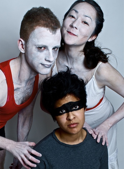 A performer in a great shirt with a black band painted around their eyes like a mask looks forward, unsettled. Behind two performers covered in white body paint stand with their hands resting on the performer in the grey. 