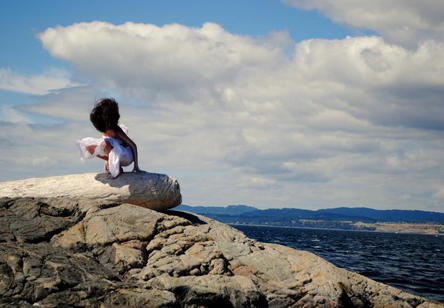 An individual dressed in white cloth squats down on a rock by the ocean. White fluffy clouds roll by overhead