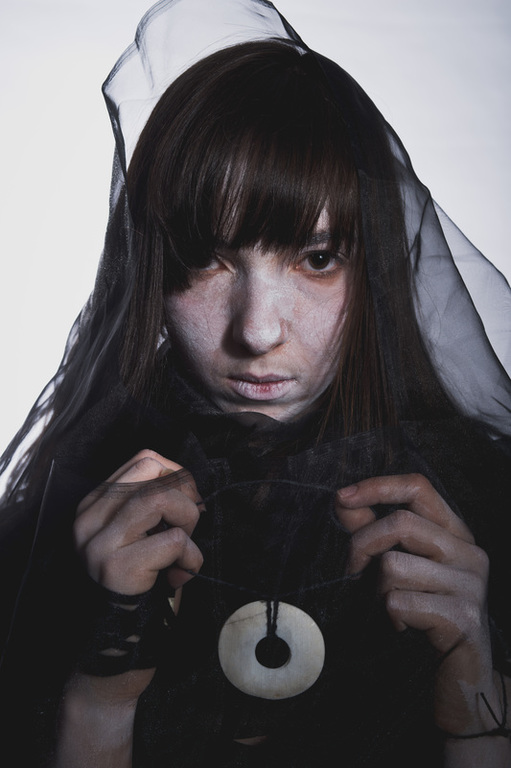 A performer with long dark hair and thick bangs stares forward. She is draped in sheer black fabric and her face and hands are painted white. She holds on to a string that is attached to a white circle with a hole in the middle. 