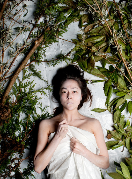Emily lays on a white background, and is wrapped in a white tarp that she grips. She stares straight forward with her lips pursed. Around her green foliage surrounds her in an arch. 