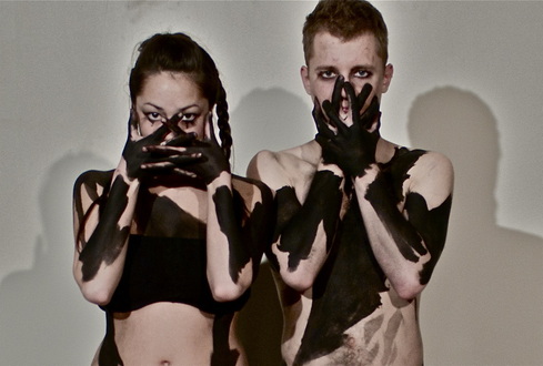 Emily and Andrew stand with their hands clasped over their mouths. They both have stripes of inky black paint running over the skin and have dark make up around their eyes