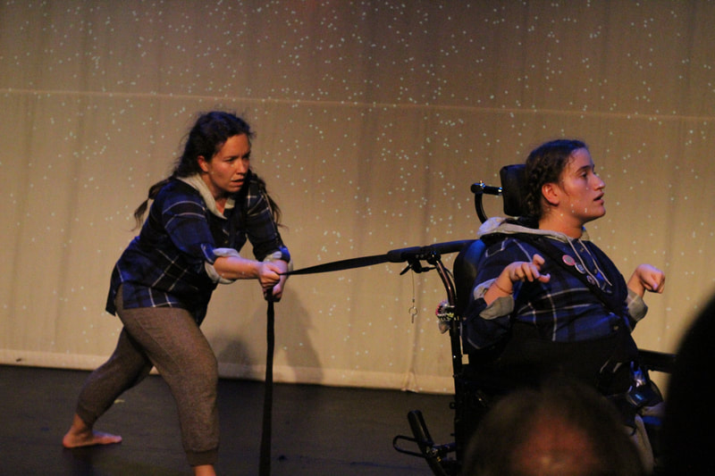 A standing dancer pulls a rope tied to the back of the other dancer’s wheelchair. 