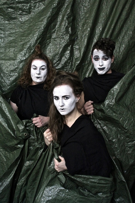 Kathleen, Randi, and Tony are holding and wrapped in the centre a large green tarp. Their faces are painted white with black accents around their eyes, eyebrows and mouth.