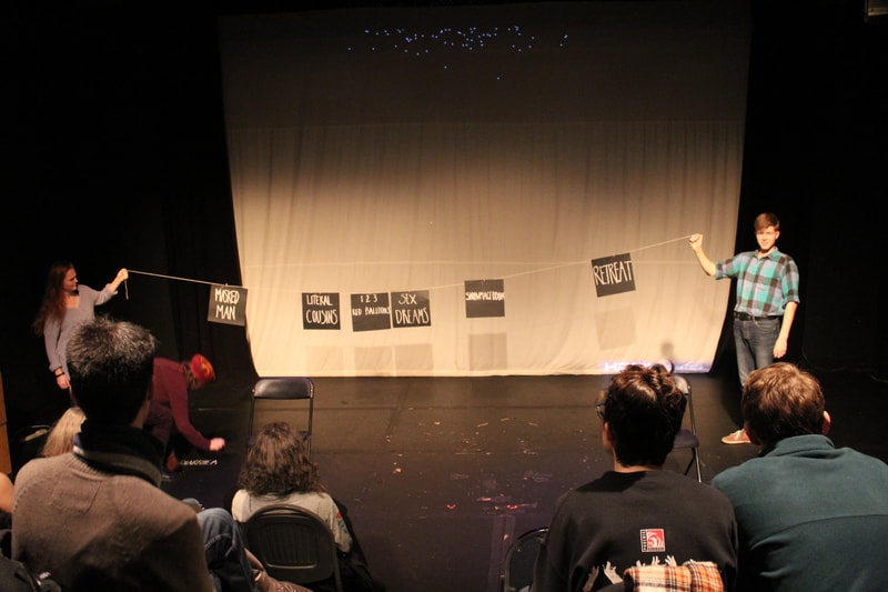 Three artists on a stage, with the backs of audience heads visible in the shot. Two of the artists are holding up a rope that has word cards attached to it. The third artist is picking up another word card.
