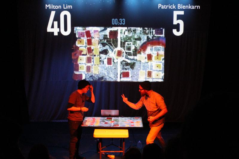Two artists stand at a table playing a game. Behind them the game and their scores are being projected onto the curtain. 