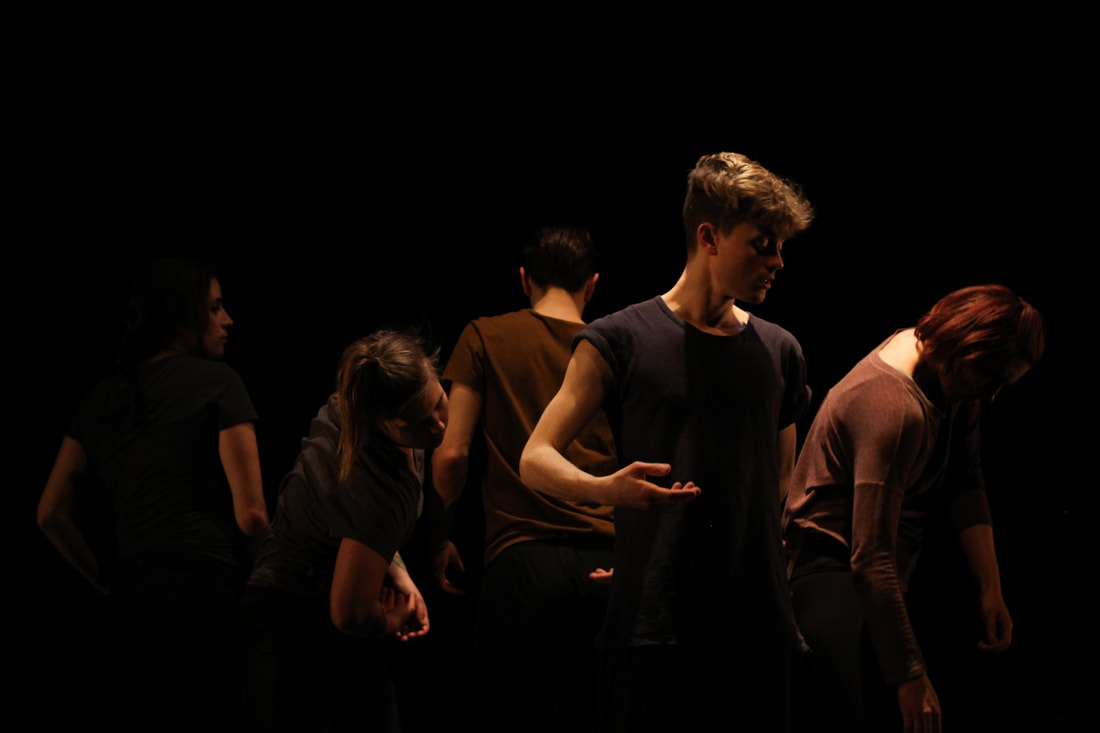 A dark image of 5 dancers in black and brown shirts standing in a small circle. They all look in different directions, none at the camera. 