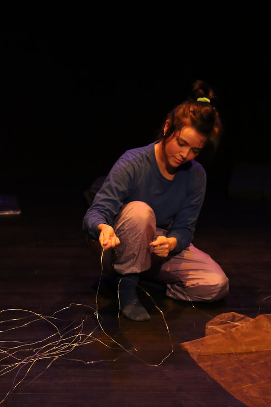 A dancer kneeling. They hold a thin piece of wire that is a attached to a pile of it on the ground beside them.