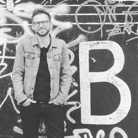 Nathan stands in front of a graffiti covered wall. He wears a jean jacket and has his hands in his pant pockets. His shoulders are slight raised as he smiles.