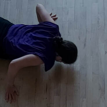 A white dancer is face down. Their arms are spread pushing on the wooden floor, and wrinkles of purple fabric are draped on their back.